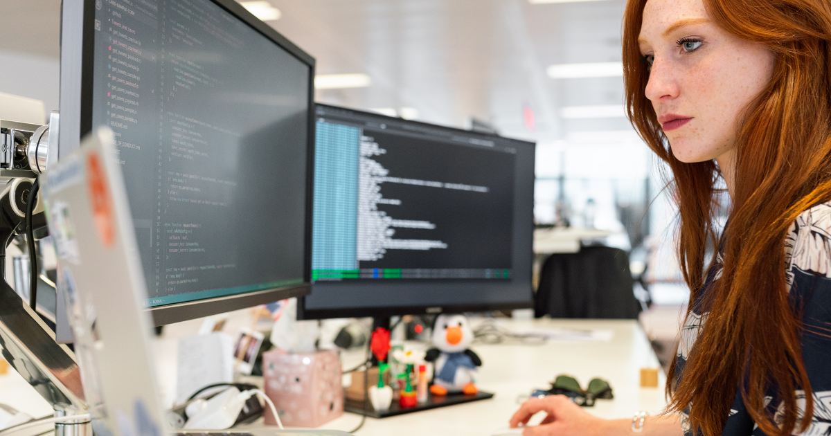 Desenvolvedora analisando linhas de código em dois monitores em um escritório